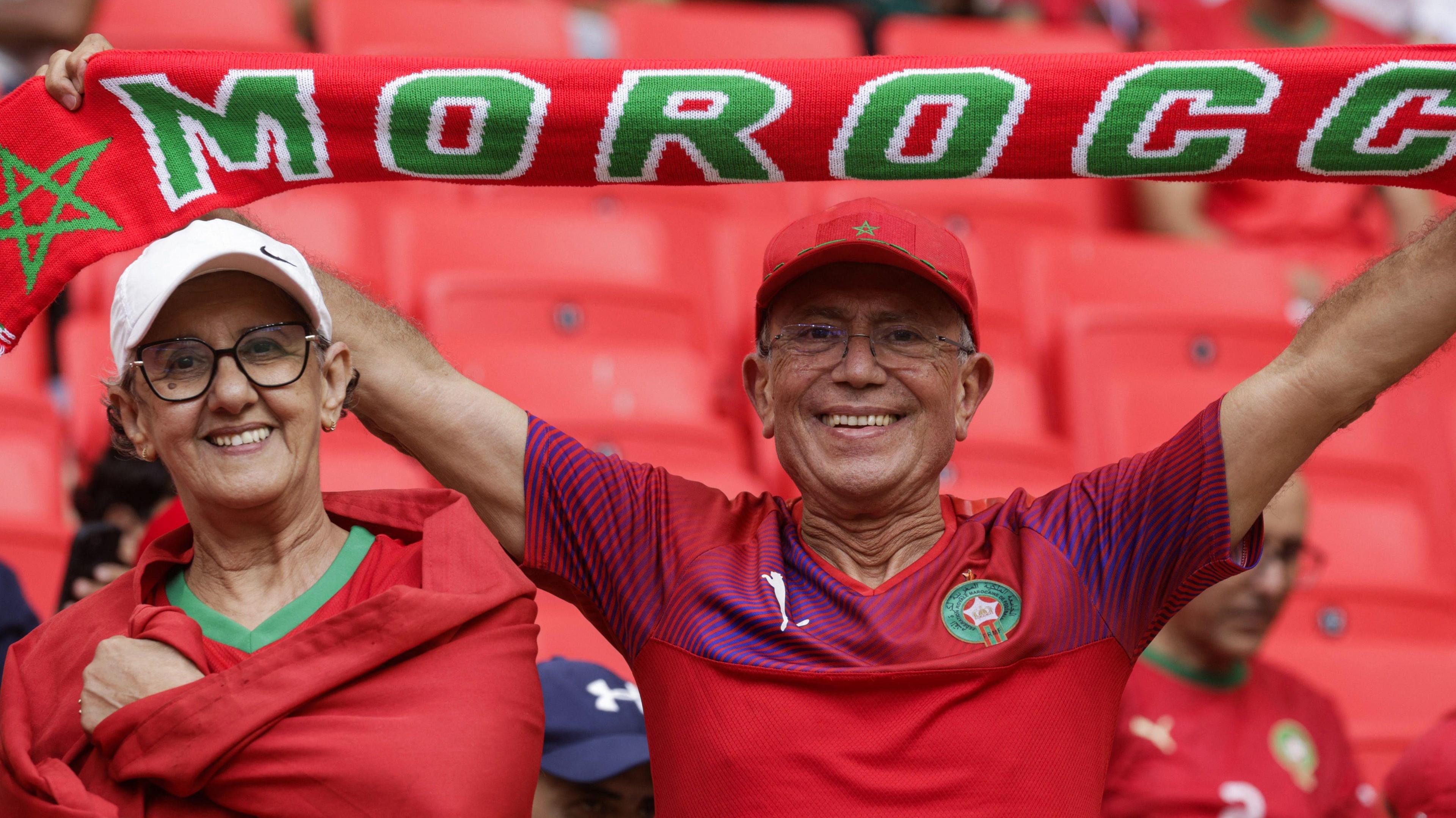 Two Morocco fans shown from chest up, dressed in red national team shirts and baseball caps, look into the camera with smiles on their faces. One holds a Morocco scarf above his head. The background behind them is a stand full of empty red seats in their folded position